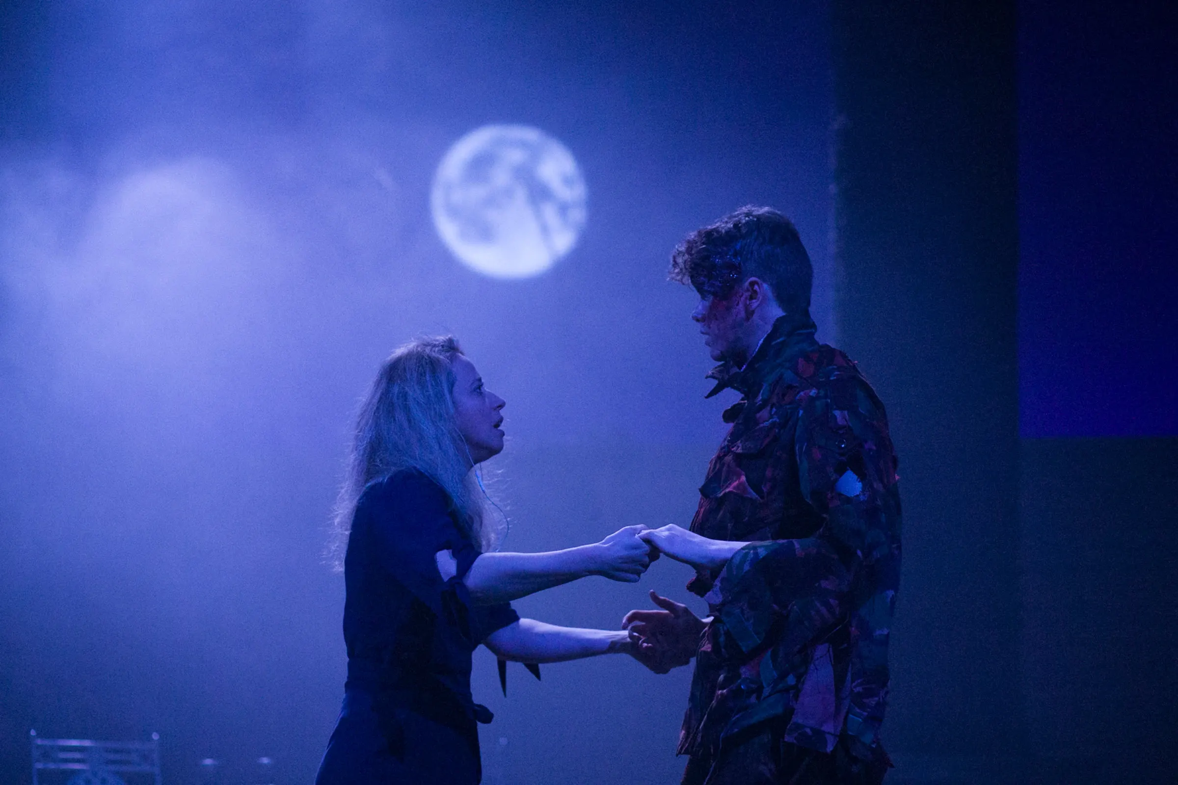 Two actors reach toward one another under blue stage lighting during a dress rehearsal of Albion