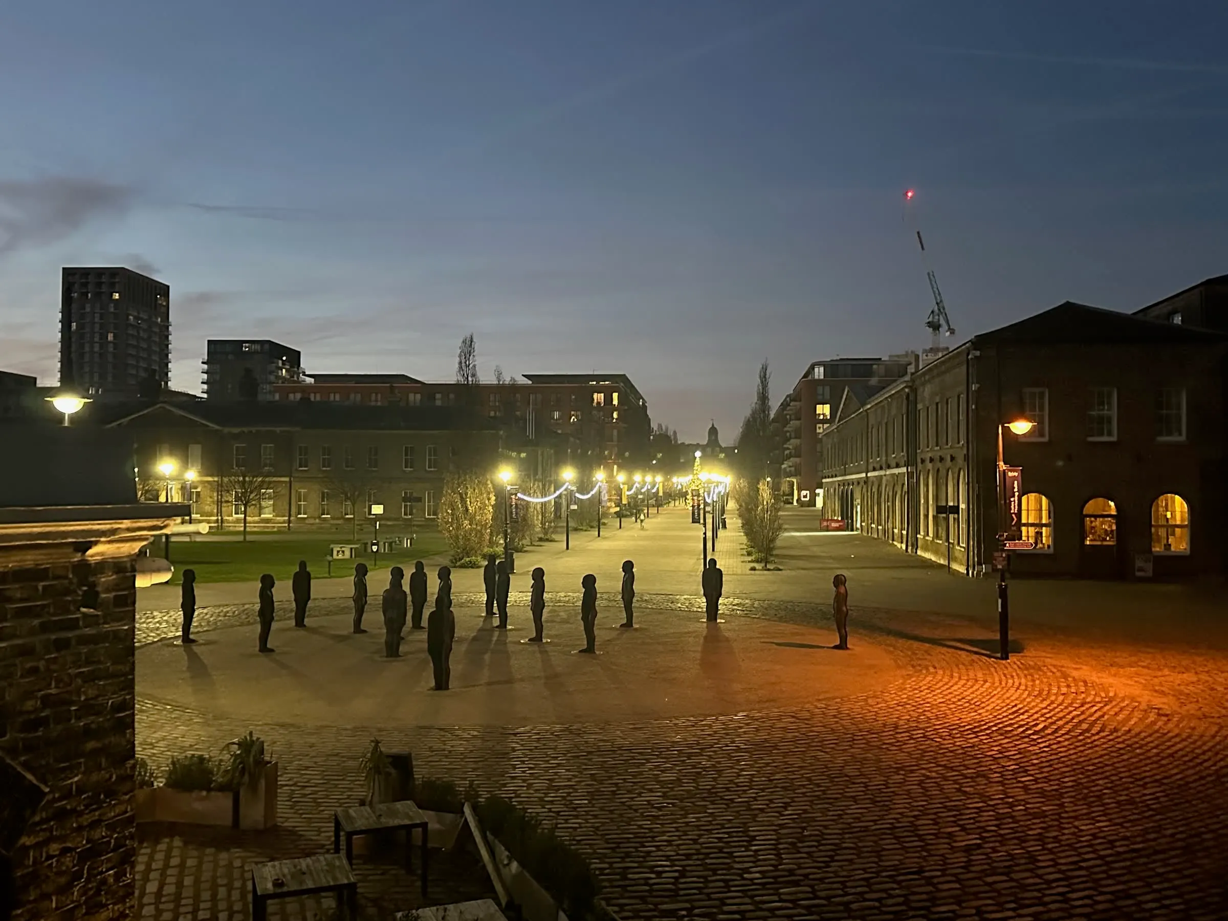 Human figures standing across a large square in Woolwich before dawn, with an illuminated avenue beyond